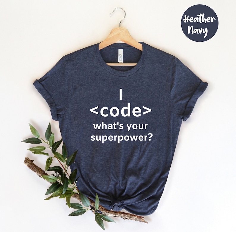 May include: Heather navy blue t-shirt with the text "I <code/> what's your superpower?" in white. The shirt is hanging on a wooden hanger. A sprig of greenery is placed to the left of the shirt.