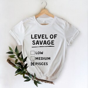 Op de afbeelding: Wit T-shirt met de zwarte tekst "LEVEL OF SAVAGE", met selectievakjes voor "LOW", "MEDIUM" en "PISCES" gemarkeerd. Het shirt hangt aan een houten hanger tegen een witte achtergrond, met groene bladeren in de linker benedenhoek.