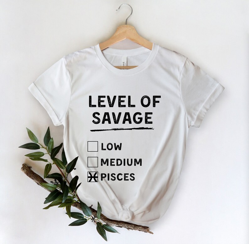Op de afbeelding: Wit T-shirt met de zwarte tekst "LEVEL OF SAVAGE", met selectievakjes voor "LOW", "MEDIUM" en "PISCES" gemarkeerd. Het shirt hangt aan een houten hanger tegen een witte achtergrond, met groene bladeren in de linker benedenhoek.