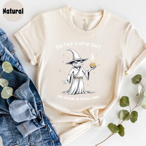 May include: A cream-coloured t-shirt featuring a cartoon wizard frog holding a fireball. The text on the shirt reads "YOU TALK A LOT OF SHIT FOR SOMEONE IN FIREBALL RANGE."
