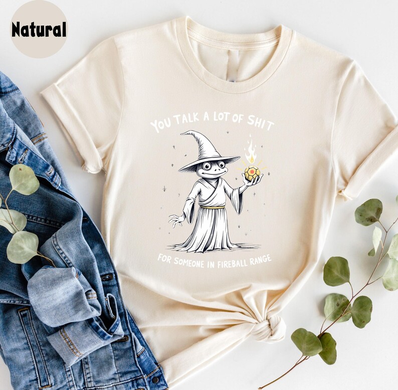 May include: A cream-coloured t-shirt featuring a cartoon wizard frog holding a fireball. The text on the shirt reads "YOU TALK A LOT OF SHIT FOR SOMEONE IN FIREBALL RANGE."