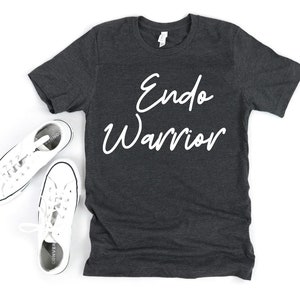 May include: Dark gray t-shirt with the words "Endo Warrior" in white script. The shirt is displayed with a pair of white canvas sneakers with black trim and laces. The sneakers are placed on a white surface.