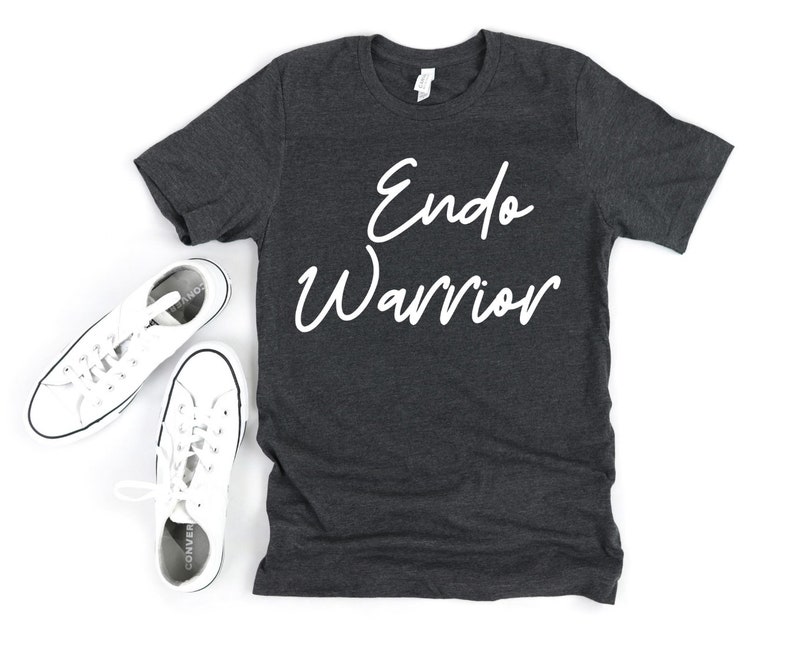 May include: Dark gray t-shirt with the words "Endo Warrior" in white script. The shirt is displayed with a pair of white canvas sneakers with black trim and laces. The sneakers are placed on a white surface.