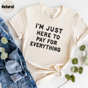 K&ouml;nnte beinhalten: Ein cremefarbenes T-Shirt mit dem Aufdruck "I'm just here to pay for everything" in Schwarz.
