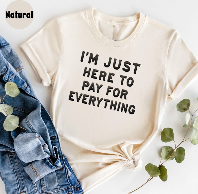 K&ouml;nnte beinhalten: Ein cremefarbenes T-Shirt mit dem Aufdruck "I'm just here to pay for everything" in Schwarz.
