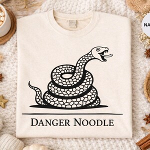 Peut inclure: Sweat-shirt beige avec un motif noir d'un serpent enroul&eacute;, la langue sortie. Le texte "DANGER NOODLE" est imprim&eacute; en dessous. Le sweat-shirt est pr&eacute;sent&eacute; sur une surface blanche textur&eacute;e avec des &eacute;l&eacute;ments d&eacute;coratifs.