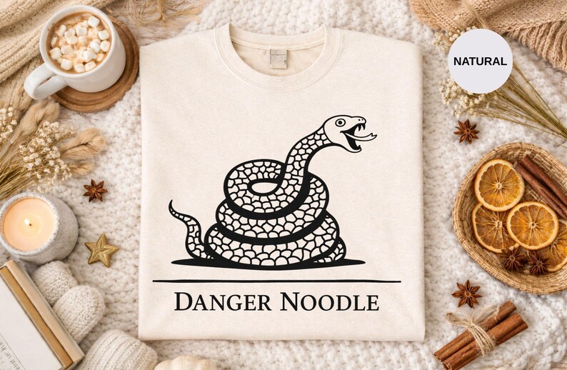 Peut inclure: Sweat-shirt beige avec un motif noir d'un serpent enroul&eacute;, la langue sortie. Le texte "DANGER NOODLE" est imprim&eacute; en dessous. Le sweat-shirt est pr&eacute;sent&eacute; sur une surface blanche textur&eacute;e avec des &eacute;l&eacute;ments d&eacute;coratifs.