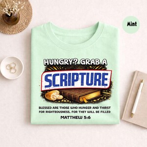 May include: Mint green t-shirt with the text "HUNGRY? GRAB A SCRIPTURE" in a bold, stylized font. Below the text is an illustration of a Bible and bread. The shirt also includes the text "BLESSED ARE THOSE WHO HUNGER AND THIRST FOR RIGHTEOUSNESS, FOR THEY WILL BE FILLED MATTHEW 5:6."