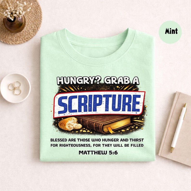May include: Mint green t-shirt with the text "HUNGRY? GRAB A SCRIPTURE" in a bold, stylized font. Below the text is an illustration of a Bible and bread. The shirt also includes the text "BLESSED ARE THOSE WHO HUNGER AND THIRST FOR RIGHTEOUSNESS, FOR THEY WILL BE FILLED MATTHEW 5:6."