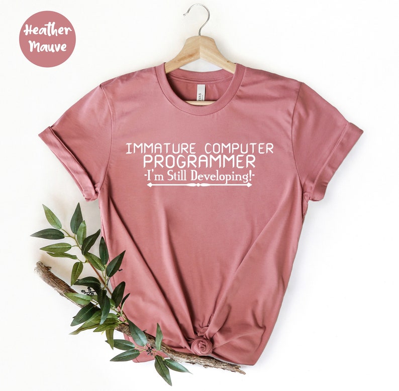 May include: Mauve t-shirt with the white text "IMMATURE COMPUTER PROGRAMMER - I'm Still Developing!". The t-shirt is displayed on a wooden hanger, with a sprig of greenery. The shirt is knotted at the bottom.