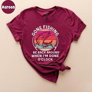 May include: Maroon t-shirt with the text "GONE FISHING" above a graphic of a fish jumping out of the water. Below the graphic, the text reads "BE BACK AROUND WHEN I'M DONE O'CLOCK".
