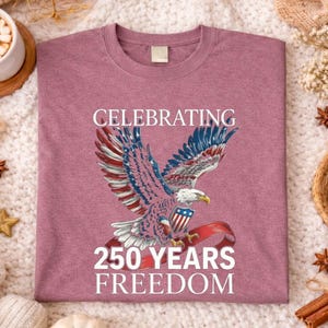 May include: Mauve-colored t-shirt featuring a patriotic eagle design with red, white, and blue accents. The text reads "CELEBRATING 250 YEARS FREEDOM". The shirt is displayed on a cozy, textured surface with decorative elements.
