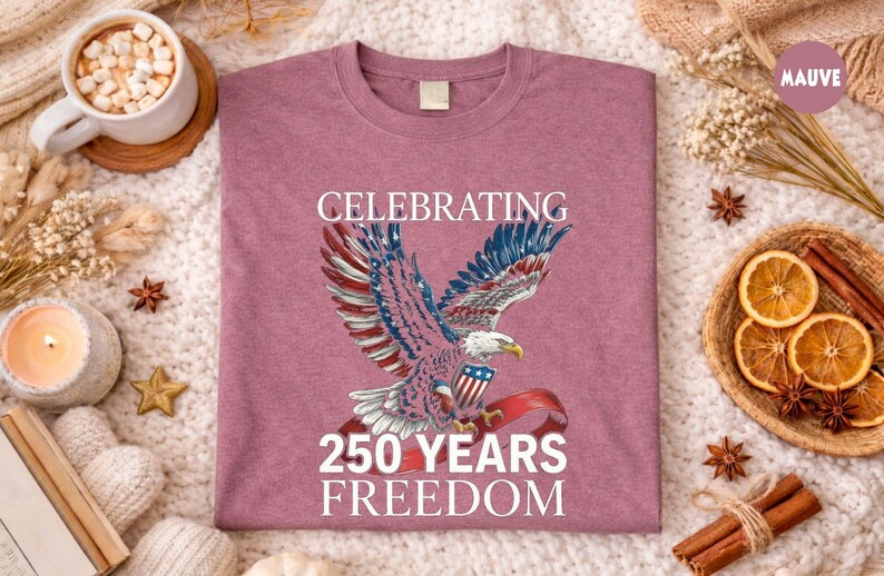 May include: Mauve-colored t-shirt featuring a patriotic eagle design with red, white, and blue accents. The text reads "CELEBRATING 250 YEARS FREEDOM". The shirt is displayed on a cozy, textured surface with decorative elements.