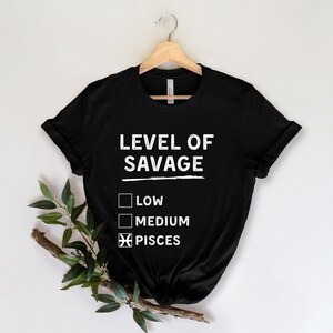 Op de afbeelding: Zwart t-shirt met de tekst "LEVEL OF SAVAGE" in het wit, met selectievakjes voor "LOW", "MEDIUM" en "PISCES" geselecteerd. Het shirt hangt aan een houten hanger tegen een witte achtergrond.