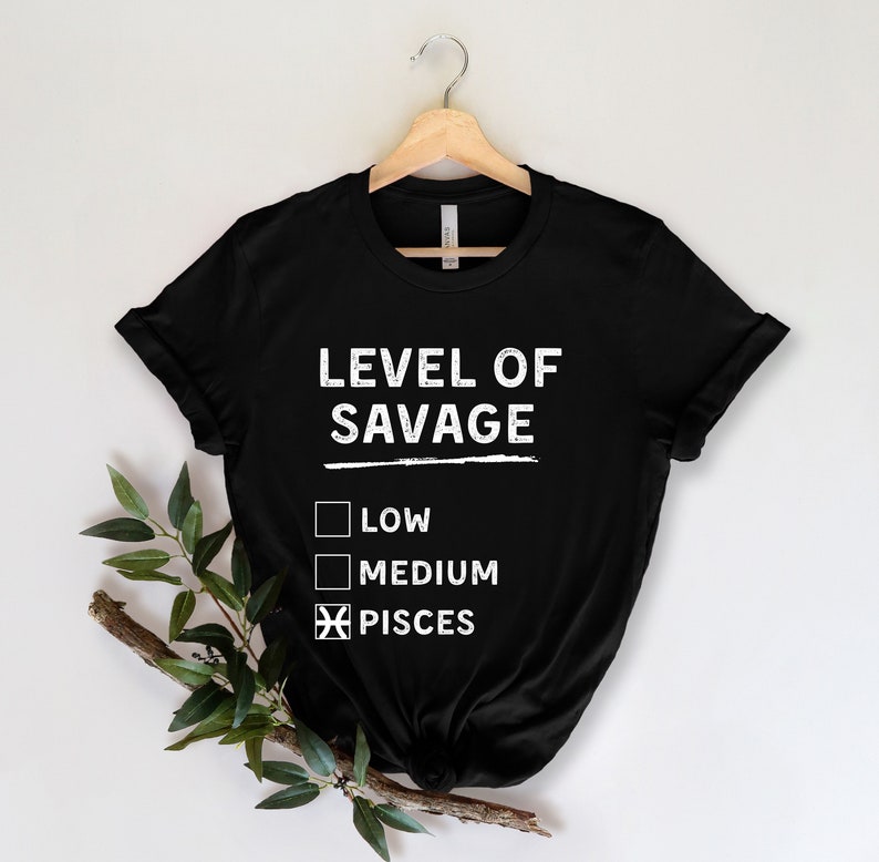 Op de afbeelding: Zwart t-shirt met de tekst "LEVEL OF SAVAGE" in het wit, met selectievakjes voor "LOW", "MEDIUM" en "PISCES" geselecteerd. Het shirt hangt aan een houten hanger tegen een witte achtergrond.