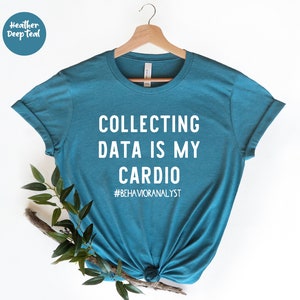 以下が含まれることがあります： 「COLLECTING DATA IS MY CARDIO」の白文字と「#BEHAVIORANALYST」のハッシュタグが入った、ティール色のTシャツ。白い背景に木製のハンガーにかけられています。左下には緑の小枝が添えられています。
