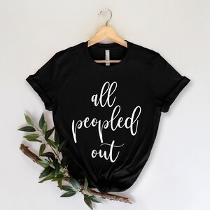 May include: Black t-shirt with the white text "all peopled out" in a cursive font. The shirt is hanging on a wooden hanger against a white background. A sprig of greenery is placed on the lower left.