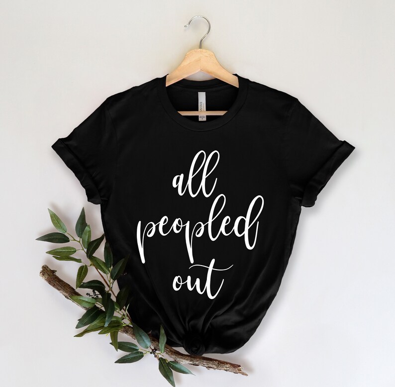 May include: Black t-shirt with the white text "all peopled out" in a cursive font. The shirt is hanging on a wooden hanger against a white background. A sprig of greenery is placed on the lower left.