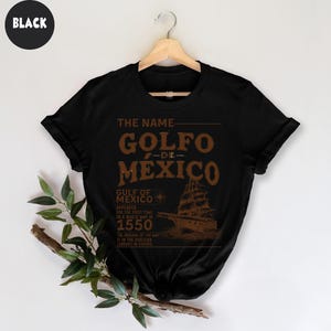 Op de afbeelding: Zwart T-shirt met een bruin grafisch ontwerp met de tekst "The Name Golfo de Mexico". De grafiek bevat ook een kompas en een schip met de tekst "Gulf of Mexico Appeared for the first time on a world map in 1550. The original of the map is in the Bodleian Library in Oxford."