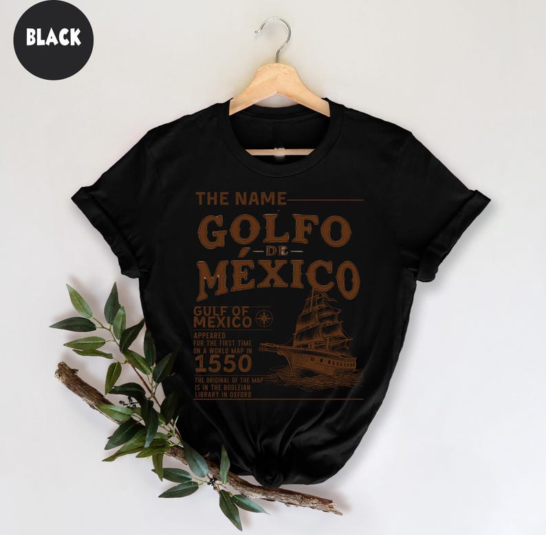 Op de afbeelding: Zwart T-shirt met een bruin grafisch ontwerp met de tekst "The Name Golfo de Mexico". De grafiek bevat ook een kompas en een schip met de tekst "Gulf of Mexico Appeared for the first time on a world map in 1550. The original of the map is in the Bodleian Library in Oxford."