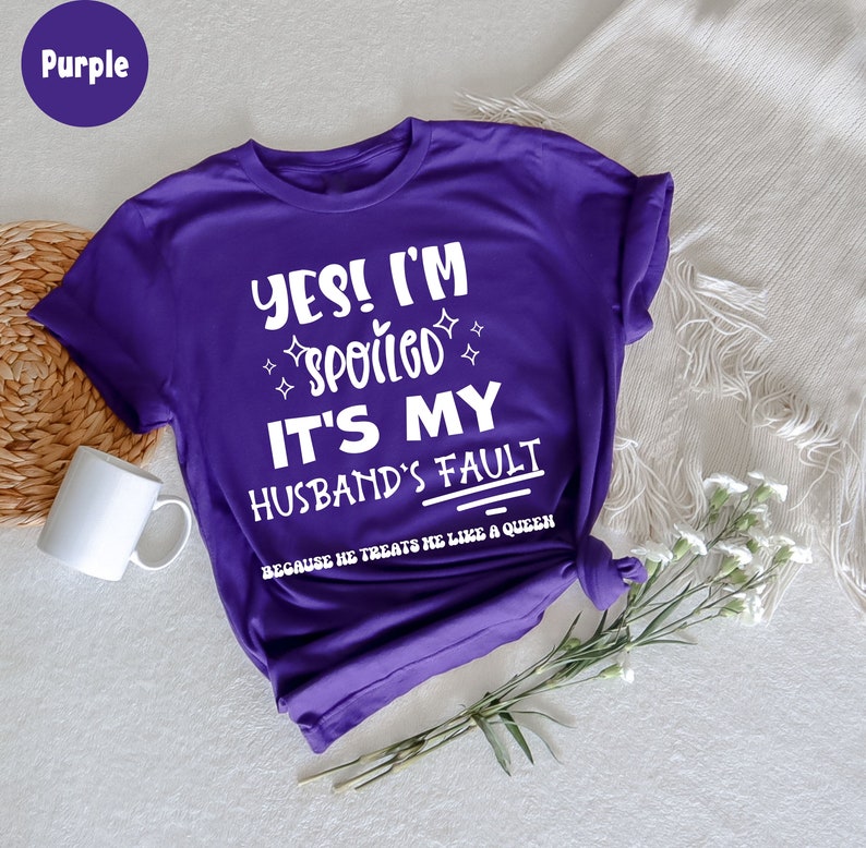 May include: Purple t-shirt with white text that reads "YES! I'M SPOILED IT'S MY HUSBAND'S FAULT BECAUSE HE TREATS ME LIKE A QUEEN"