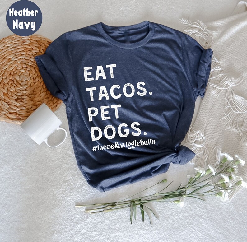 Op de afbeelding: Een heideblauwe t-shirt met de tekst "EAT TACOS. PET DOGS. #tacos&wigglebutts" in wit gedrukt.