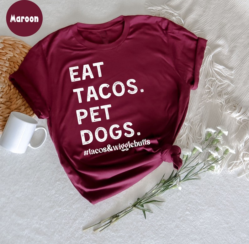 Op de afbeelding: Een maroon t-shirt met witte tekst die "Eat Tacos. Pet Dogs. #tacos&wigglebutts" zegt.