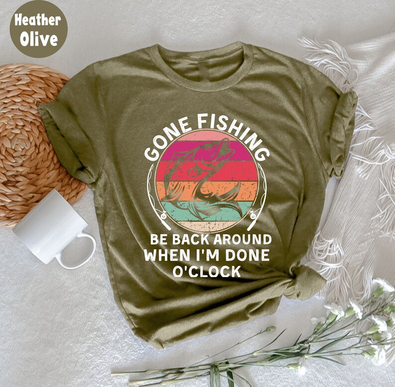 May include: Heather olive green t-shirt with a circular graphic that reads "GONE FISHING" above a fish illustration. The text "BE BACK AROUND WHEN I'M DONE O'CLOCK" is below the graphic. The shirt is folded and tied at the bottom.