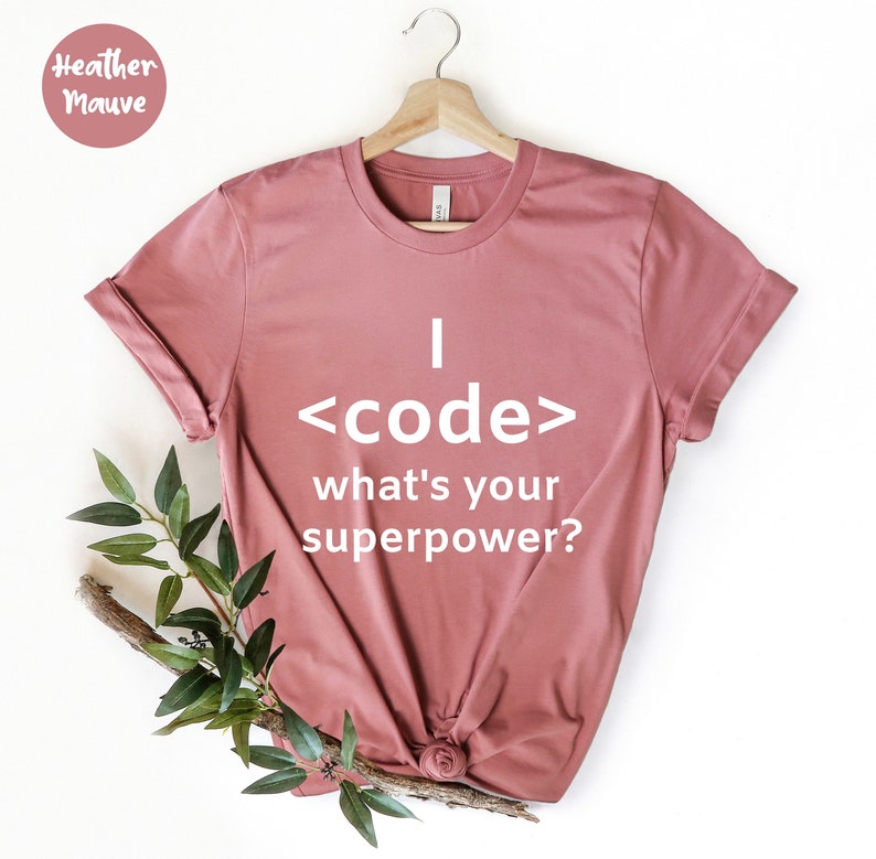 May include: Heather mauve t-shirt with the text "I <code/> what's your superpower?" in white. The shirt is displayed on a wooden hanger with a sprig of greenery.