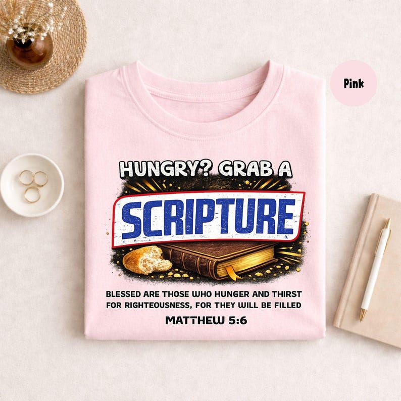 May include: Pink t-shirt with the text "HUNGRY? GRAB A SCRIPTURE" in a bold, stylized font. Below the text is an illustration of a loaf of bread and a book. The text "BLESSED ARE THOSE WHO HUNGER AND THIRST FOR RIGHTEOUSNESS, FOR THEY WILL BE FILLED MATTHEW 5:6" is also printed on the shirt.