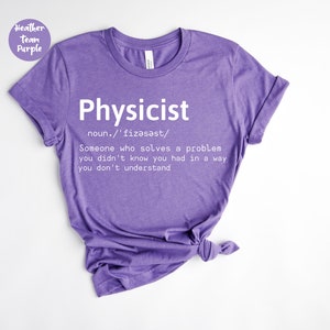 Könnte beinhalten: Ein lilafarbenes T-Shirt mit dem Wort "Physicist" in Weiß aufgedruckt. Die Definition von Physiker ist ebenfalls auf dem T-Shirt abgedruckt. Das T-Shirt besteht aus einem weichen Material und hat kurze Ärmel.