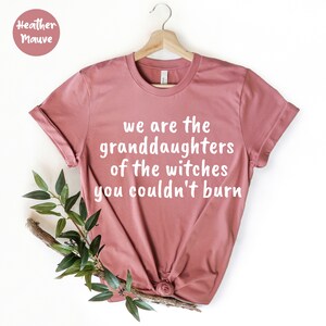 May include: Heather mauve t-shirt with the text "we are the granddaughters of the witches you couldn't burn" in white. The shirt is displayed on a wooden hanger with a sprig of greenery.
