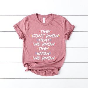 May include: A pink t-shirt with the text "They don't know that we know they know we know" in white with colorful dots.