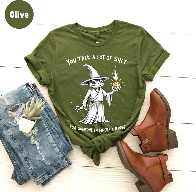 May include: Olive green t-shirt with the text "You Talk A Lot Of Shit For Someone In Fireball Range" and a cartoon wizard frog graphic. The image also shows blue jeans, brown ankle boots, and a small plant.