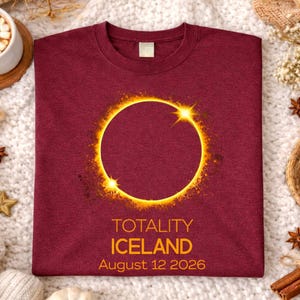 Op de afbeelding: Bordeauxrood T-shirt met een gouden zonsverduistering ontwerp. De tekst "TOTALITY ICELAND August 12 2026" is eronder gedrukt. Het shirt wordt gepresenteerd op een witte, getextureerde ondergrond met decoratieve elementen.