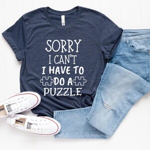 Pode incluir: Uma camiseta azul marinho com o texto branco "SORRY I CAN'T I HAVE TO DO A PUZZLE". A camiseta &eacute; combinada com jeans azul claro e t&ecirc;nis Converse brancos. O conjunto est&aacute; sobre uma superf&iacute;cie branca.