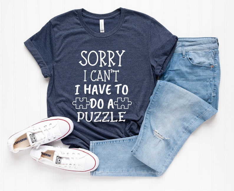 Pode incluir: Uma camiseta azul marinho com o texto branco "SORRY I CAN'T I HAVE TO DO A PUZZLE". A camiseta &eacute; combinada com jeans azul claro e t&ecirc;nis Converse brancos. O conjunto est&aacute; sobre uma superf&iacute;cie branca.