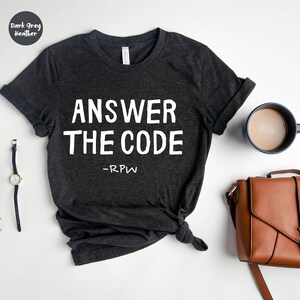 Könnte beinhalten: Dunkelgraues meliertes T-Shirt mit dem weißen Text "ANSWER THE CODE" und "-RPW" darunter. Das Shirt ist mit hochgekrempelten Ärmeln und einem Knoten am Saum versehen. Eine Uhr, eine Kaffeetasse und eine braune Handtasche sind ebenfalls im Bild.