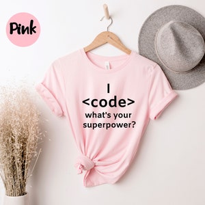 May include: A light pink t-shirt with the text "I <code/> what's your superpower?" in black. The shirt is hanging on a wooden hanger. A pink circle with the word "Pink" is in the upper left corner.