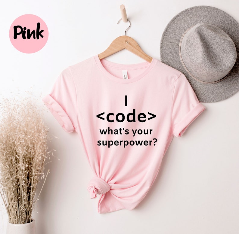 May include: A light pink t-shirt with the text "I <code/> what's your superpower?" in black. The shirt is hanging on a wooden hanger. A pink circle with the word "Pink" is in the upper left corner.