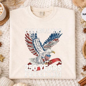 May include: Cream-colored sweatshirt featuring a patriotic eagle design with red, white, and blue accents. The text reads "CELEBRATING 250 YEARS FREEDOM". The design is centered on the front of the shirt.