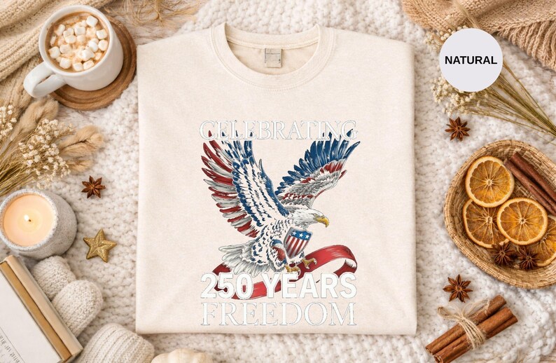 May include: Cream-colored sweatshirt featuring a patriotic eagle design with red, white, and blue accents. The text reads "CELEBRATING 250 YEARS FREEDOM". The design is centered on the front of the shirt.