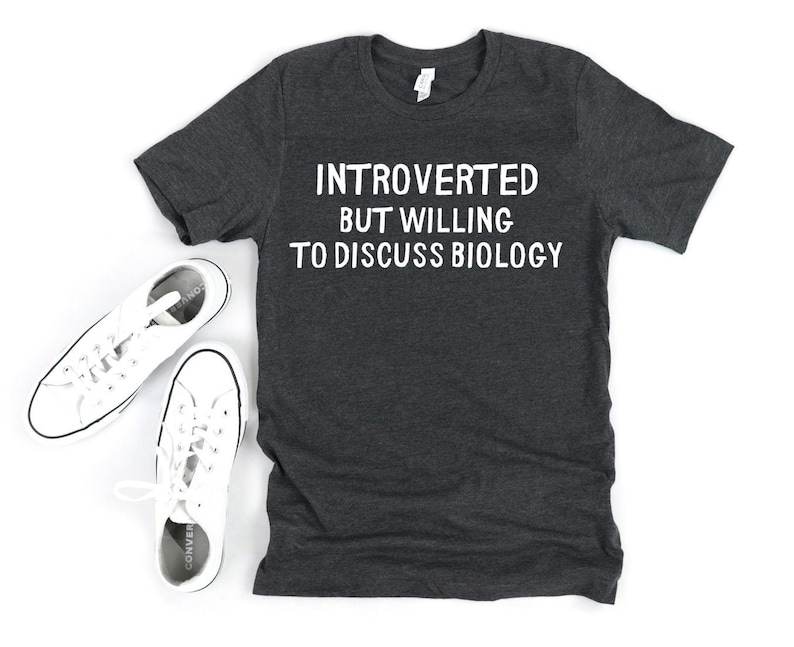 May include: Dark gray t-shirt with the text "INTROVERTED BUT WILLING TO DISCUSS BIOLOGY" in white. Two pairs of white canvas sneakers are next to the shirt on a white surface.