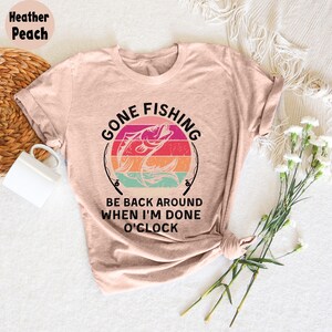 May include: Heather peach t-shirt with a fishing-themed graphic. The design features a fish jumping out of the water, with the words "GONE FISHING" and "BE BACK AROUND WHEN I'M DONE O'CLOCK" in black text. The shirt is styled with rolled sleeves.