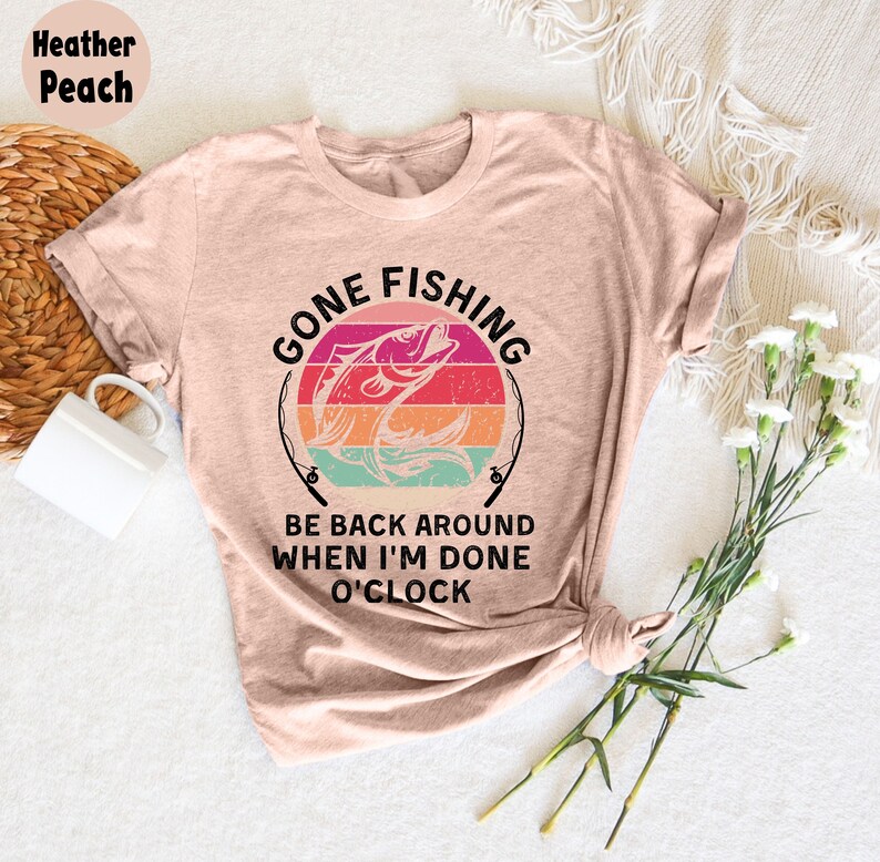 May include: Heather peach t-shirt with a fishing-themed graphic. The design features a fish jumping out of the water, with the words "GONE FISHING" and "BE BACK AROUND WHEN I'M DONE O'CLOCK" in black text. The shirt is styled with rolled sleeves.