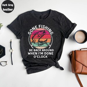 May include: Dark grey heather t-shirt with a colorful graphic. The graphic reads "GONE FISHING" above a leaping fish design, with the text "BE BACK AROUND WHEN I'M DONE O'CLOCK" below. The shirt is knotted at the bottom.