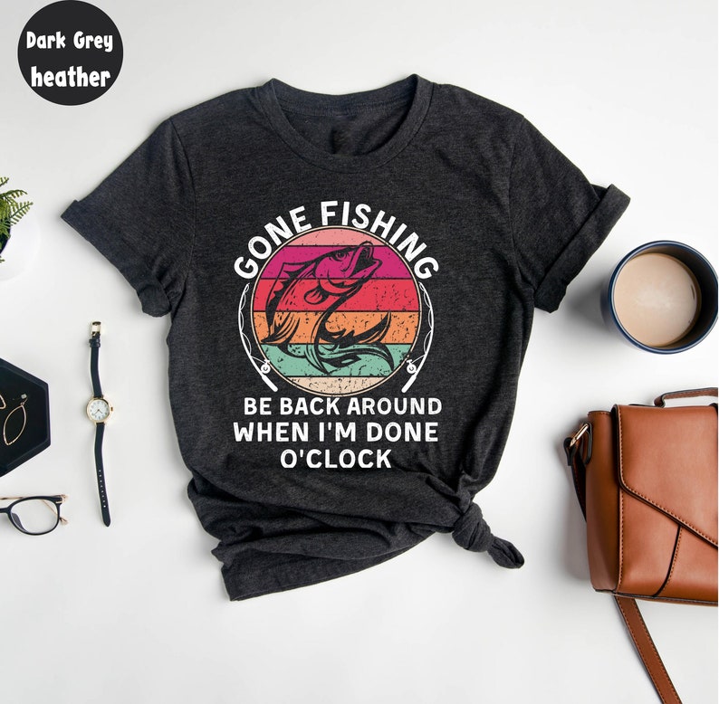 May include: Dark grey heather t-shirt with a colorful graphic. The graphic reads "GONE FISHING" above a leaping fish design, with the text "BE BACK AROUND WHEN I'M DONE O'CLOCK" below. The shirt is knotted at the bottom.