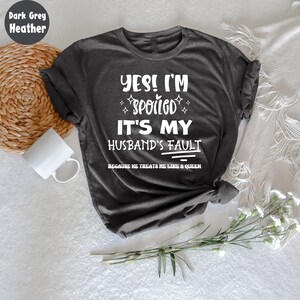 May include: Dark grey heather t-shirt with white text that reads "YES! I'M SPOILED IT'S MY HUSBAND'S FAULT BECAUSE HE TREATS ME LIKE A QUEEN".
