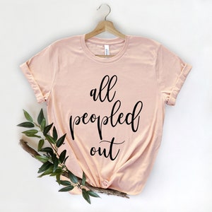 May include: A light pink t-shirt with rolled-up sleeves, hanging on a wooden hanger. The shirt features the phrase "all peopled out" in black cursive script. A sprig of greenery is placed on the lower left side of the shirt.