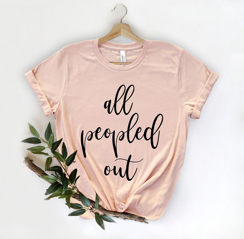 May include: A light pink t-shirt with rolled-up sleeves, hanging on a wooden hanger. The shirt features the phrase "all peopled out" in black cursive script. A sprig of greenery is placed on the lower left side of the shirt.